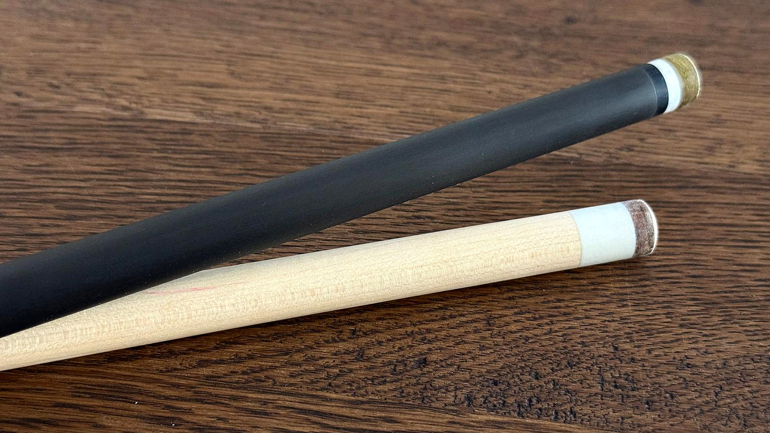 A natural Canadian hard rock maple pool cue shaft with white ferrule and leather tip lying next to a matte black carbon fiber shaft with brass ferrule on a dark oak table